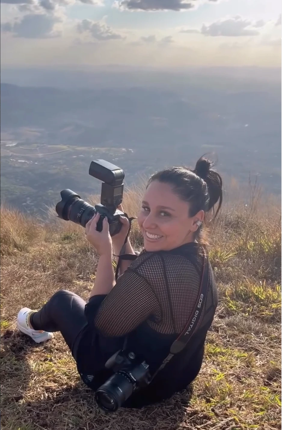Lu Prado sorrindo com a câmera na mão, sentada no alto de uma serra